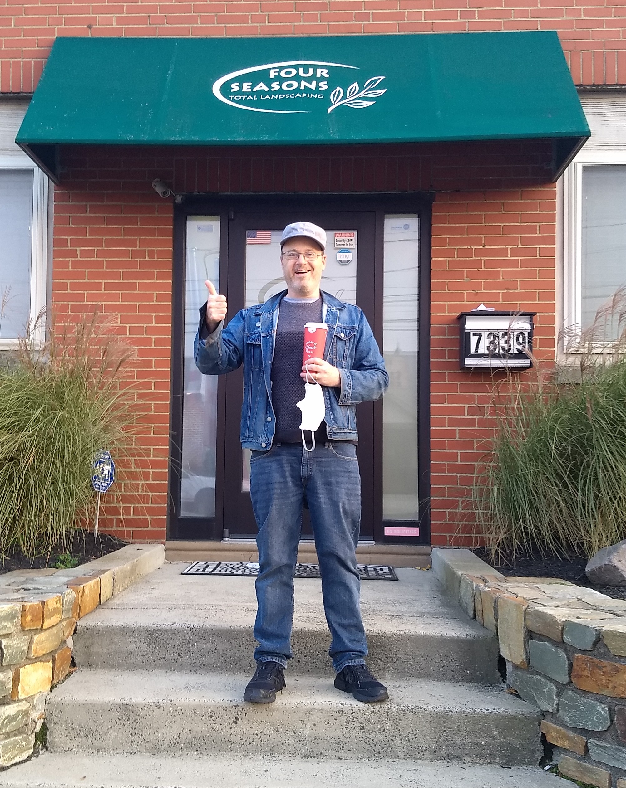 I am smiling in front of the main entrance of Four Seasons Total
Landscaping in the Tacony neighborhood of Philadelphia.  I am wearing
a gray cap and sweater, jeans, and a blue denim jacket.  I have a red
paper Wawa coffee cup in one hand, and am giving a thumbs up with the
other.